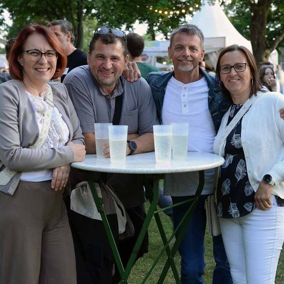 Vier Personen posieren an einem Tisch mit Gläsern Wasser. Zwei Frauen und zwei Männer. Bäume und Zelte im Hintergrund.