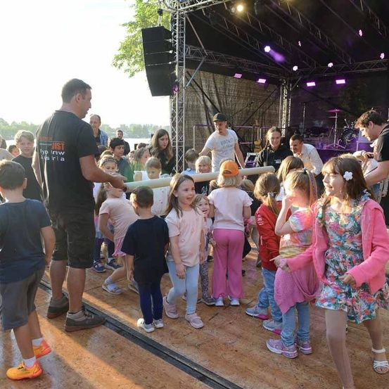 Eine Gruppe von Kindern und Erwachsenen steht auf einer Bühne. Einige Kinder halten einen langen Stab. Ein erwachsener Mann steht vor ihnen. Hinter ihnen steht eine Bühne mit Lautsprechern, einem Schlagzeug und anderen Musikinstrumenten.