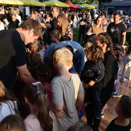 Eine Menschenmenge, bestehend aus Erwachsenen und Kindern, versammelt sich im Freien. Einige stehen, andere sitzen. Der Mann vorne lehnt sich nach unten, um die Kinder zu betrachten.