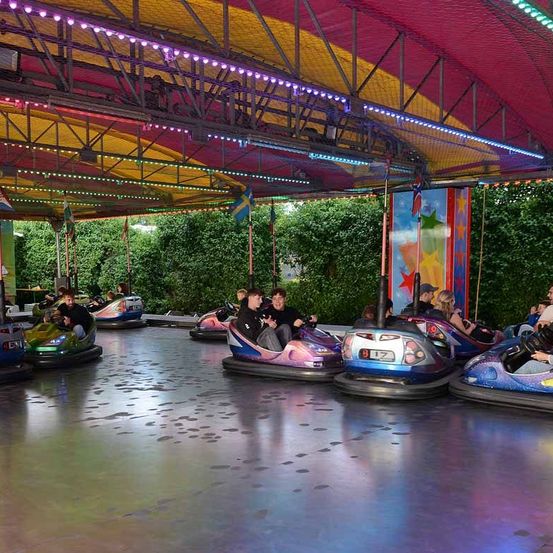 Eine Gruppe von Menschen genießt die Fahrt mit den Bumper Cars unter einem bunten Baldachin mit Flaggen und Lichtern. Die Bumper Cars stehen auf einer nassen Fläche mit Lichtreflexionen.