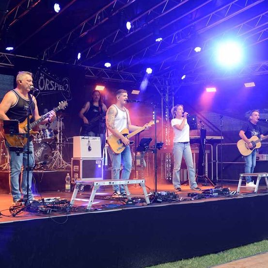 Eine Band tritt auf einer Bühne mit lila und blauen Lichtern auf. Ein Mann spielt Gitarre, während ein anderer in ein Mikrofon singt. Ein weiterer Gitarrist und Schlagzeuger sind ebenfalls auf der Bühne.