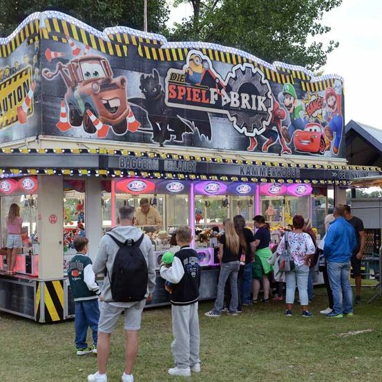 Menschen warten in einer Schlange an einem Jahrmarktstand. Er ist mit Auto- und Minion-Charakteren dekoriert. Der Stand hat Greifautomaten und ein Schild, das 'Spiel-Brik' lautet.