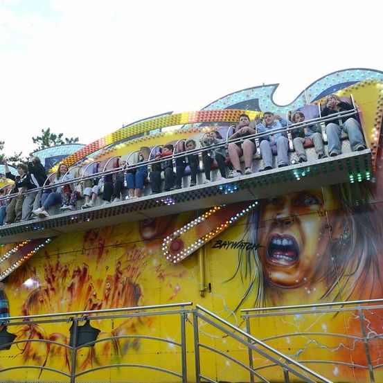 Eine Gruppe von Menschen fährt auf einem bunten Jahrmarktkarussell mit einer gelben Wand, die ein schreiendes Gesicht zeigt.