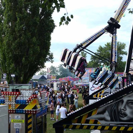 Ein lebhaftes Jahrmarktgelände mit einem bunten Baum, einem Riesenrad und einer Fahrt mit dem Bild einer Frau. Menschen sind versammelt, einige warten auf Fahrten.