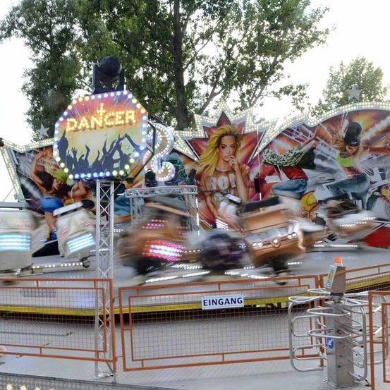 Eine sich drehende Fahrt namens 'Dancer' ist in Aktion, mit verschwommenen Figuren auf Sitzen und einem bunten Hintergrund. Der Eingang ist mit einem Schild gekennzeichnet, das 'Eingang' sagt.