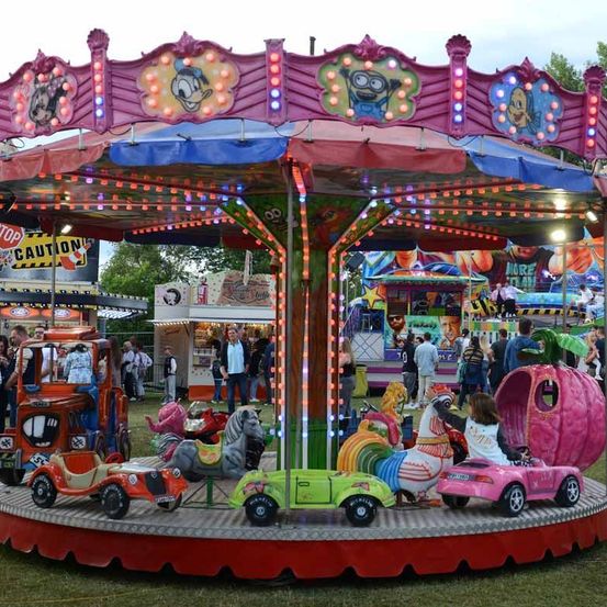 Ein farbenfroher Karussell im Freien mit Lichtern und Fahrgeschäften, darunter Autos und Pferde, umgeben von Menschen auf einem Jahrmarkt.