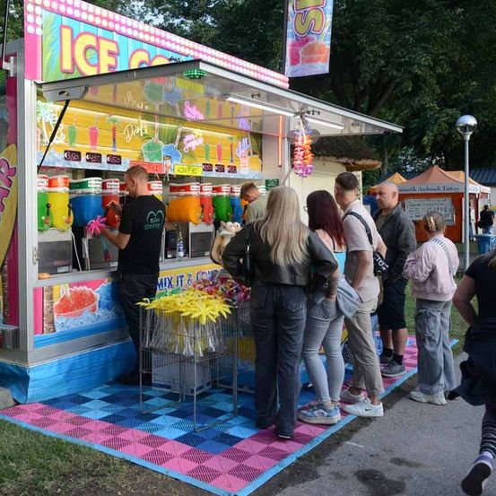 Menschen stehen in einer Schlange an einem Eiswagen mit buntem Angebot. Ein Mann in Schwarz serviert Getränke. Der Wagen hat eine Mix-it-yourself-Option.