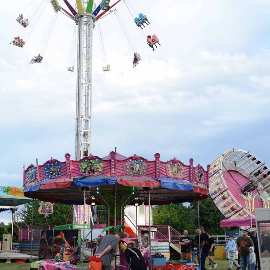 Menschen fahren auf einer bunten Karussell und einer Drehscheibe auf einem Freiluftfest. Andere Fahrgeschäfte und Attraktionen sind im Hintergrund zu sehen.