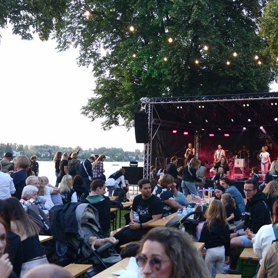 Ein Outdoor-Musikereignis mit Menschen, die vor einer Bühne mit einer auftretenden Band sitzen und stehen. Bäume und Lichter sind über ihnen.