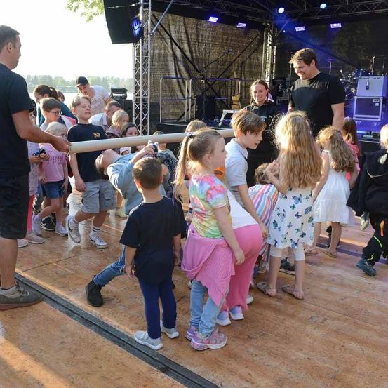 Eine Gruppe von Kindern und Erwachsenen versammelt sich auf einer Bühne, wobei zwei Männer ein langes Objekt halten. Die Bühne hat einen Holzboden, eine Kulisse und Musikinstrumente. Einige Leute schauen zur Kamera.