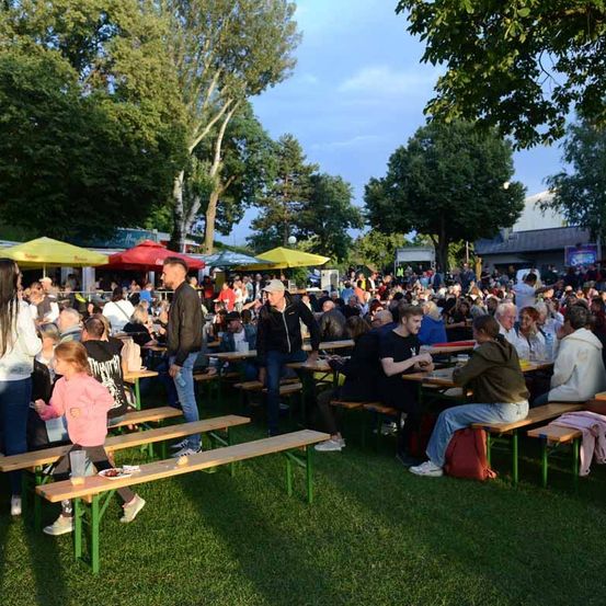 Eine große Menschenmenge versammelt sich in einem Park mit langen Holzbänken und bunten Sonnenschirmen. Viele sitzen, während andere stehen. Die Atmosphäre ist lebhaft, mit Menschen, die plaudern und Essen genießen. Die Bäume im Hintergrund deuten auf einen warmen Abend hin.