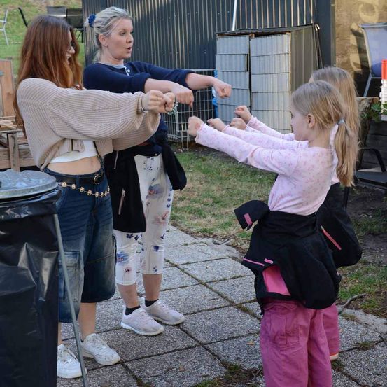Drei Frauen und zwei Kinder interagieren im Freien. Eine Frau trägt einen beigefarbenen Pullover, während eine andere in einem blauen Oberteil Karate-Bewegungen nachahmt. Ein kleines Mädchen in rosa Hosen und einer Jacke spielt mit den Kindern.