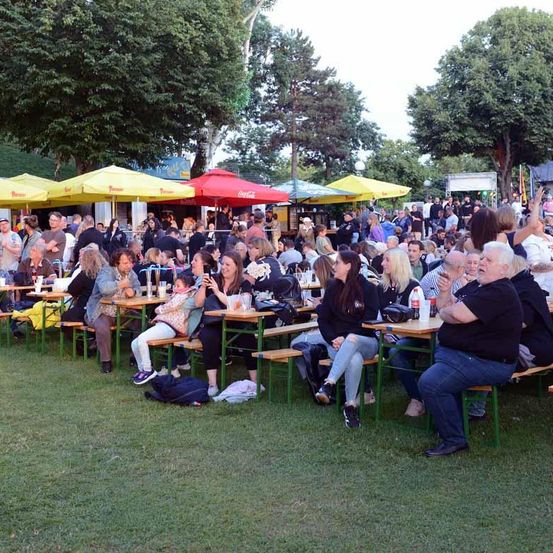 Eine Menschenmenge sitzt auf einer Rasenfläche an Tischen, unter gelben und roten Sonnenschirmen. Einige tragen Freizeitkleidung, andere sind formeller gekleidet. Bäume und ein klarer Himmel bilden den Hintergrund.