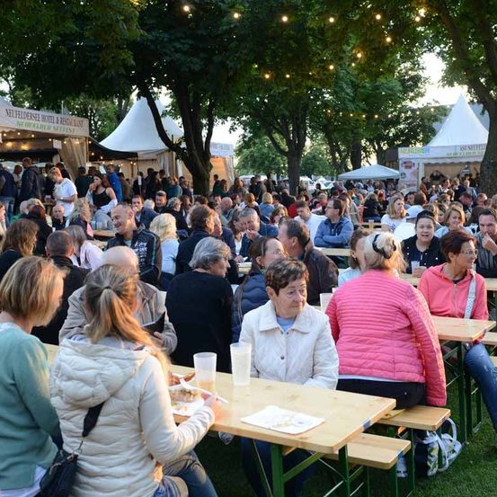 Eine große Menschenmenge sitzt auf Tischen in einem Außenbereich, möglicherweise für eine Veranstaltung. Einige Leute essen und trinken. In der Nähe befinden sich Zelte mit Bannern.