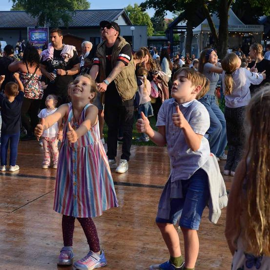 Mehrere Kinder tanzen in einem Außenbereich. Ein Mädchen in einem gestreiften Kleid lächelt in die Kamera. Ein Junge gibt einen Daumen hoch. Hinter ihnen stehen Menschen und genießen die Veranstaltung.