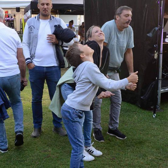 Eine Familie und zwei weitere Erwachsene befinden sich auf einem Rasenfeld, wahrscheinlich bei einer Veranstaltung. Der Junge schaut nach oben, während die Frau und der Mann neben ihm lächeln. Der Mann auf der linken Seite hält eine Jacke. Im verschwommenen Hintergrund sind Zelte, ein Lautsprecher und Menschen zu sehen.