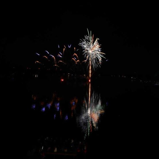 Ein nächtliches Feuerwerk bricht am Himmel hervor und erleuchtet den dunklen Himmel mit bunten Lichtern und Reflexionen auf dem ruhigen Wasser.