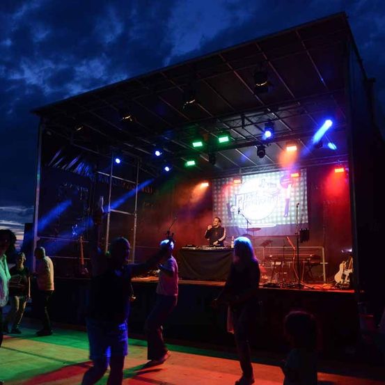 Ein nächtliches Konzert auf einer Bühne mit leuchtenden Farben und einem DJ. Vor der Bühne tanzen Menschen.