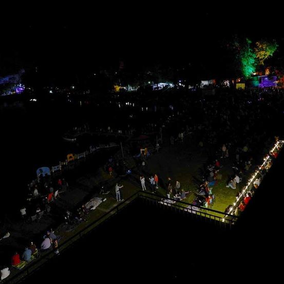 Eine Luftaufnahme eines nächtlichen Treffens mit vielen Menschen, die auf dem Rasen sitzen, einige stehen, und farbige Lichter erhellen das Gebiet, zusammen mit Bäumen und Gebäuden in der Ferne.