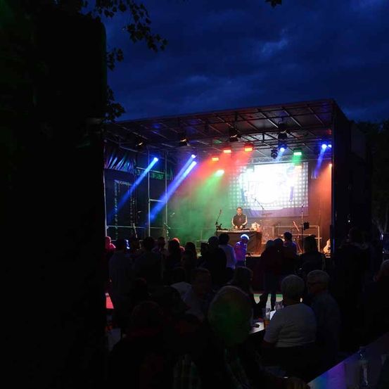 Ein nächtliches Musikevent zeigt einen DJ auf der Bühne, beleuchtet von leuchtenden, mehrfarbigen Lichtern. Ein Publikum versammelt sich davor, einige sitzend und andere stehend, unter einem teilweise bewölkten Himmel.