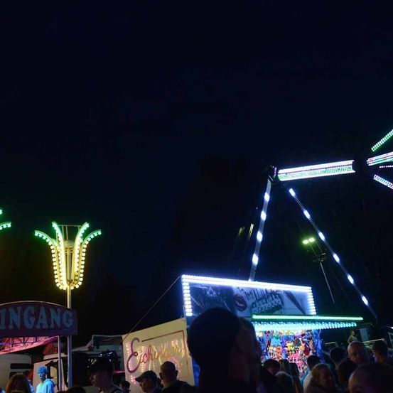 Eine Menschenmenge versammelt sich bei Nacht um eine beleuchtete Jahrmarkt-Attraktion, mit einem Schild, das 'Eingang' sagt, und einem hohen, farbenfrohen Turm mit Lichtern.