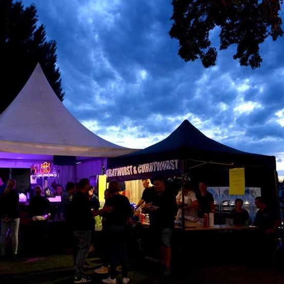Ein Outdoor-Essenstand mit violettem Licht beleuchtet eine Gruppe von Menschen, einige stehend, andere sitzend, unter einem Zelt mit blauem Baldachin.