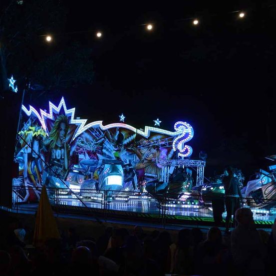 Ein nächtlicher Karussell-Fahrt mit bunten Lichtern und einer Menge, die von unten zuschaut. Ein gelber Regenschirm ist im Vordergrund sichtbar.