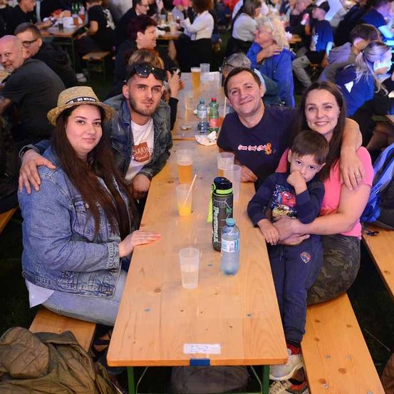 Eine Familie sitzt lächelnd an einem Tisch im Freien. Dahinter sitzen eine Gruppe von Menschen an anderen Tischen. Der Tisch hat Getränke und Essen.