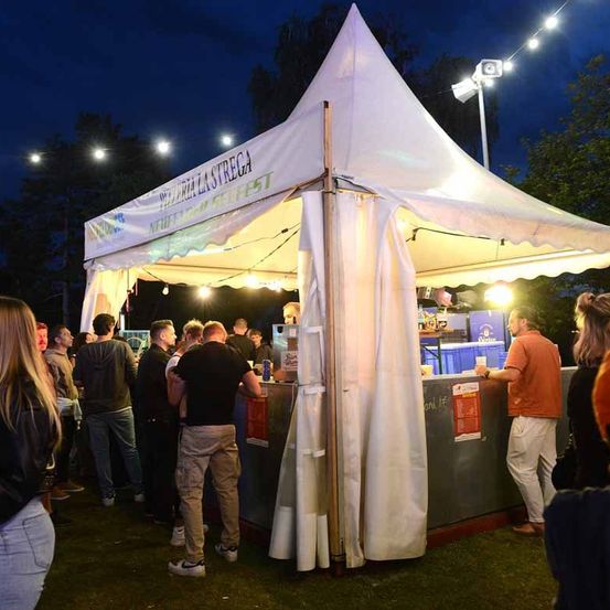 Ein Imbissstand steht unter einem weißen Zelt mit der Aufschrift "Pizzeria la Strega". Es ist Nacht und es stehen Menschen in der Schlange.