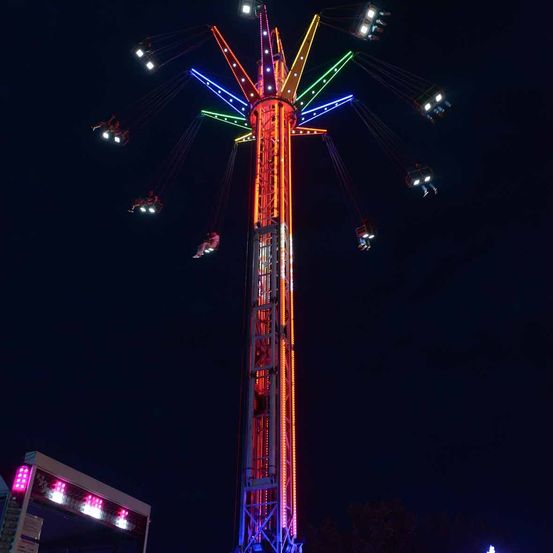 Ein hoher, bunter Riesenrad mit Lichtern ist nachts in Bewegung. Leute sitzen darauf. Die Lichter sind rot, gelb, grün und blau.
