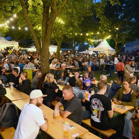Ein Outdoor-Festival mit vielen Menschen, die an Tischen sitzen, trinken und plaudern. Bäume mit Lichterketten im Hintergrund. Zelte und andere Strukturen sind in der Ferne zu sehen.