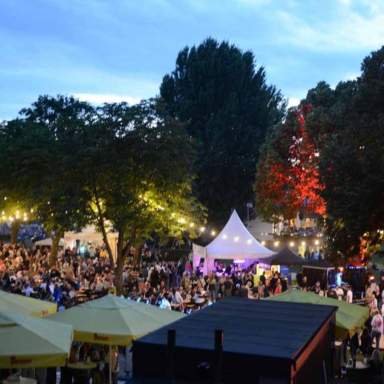 Eine nächtliche Outdoor-Veranstaltung mit vielen Menschen, Zelten und Bäumen. Die Atmosphäre ist lebhaft mit Musik und Essensständen.