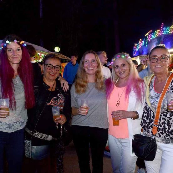 Fünf Frauen stehen bei Nacht zusammen, lächeln und halten Getränke. Hinter ihnen ist eine bunte Lichtshow zu sehen, und mehrere Personen sind im Hintergrund, möglicherweise bei einem Festival.