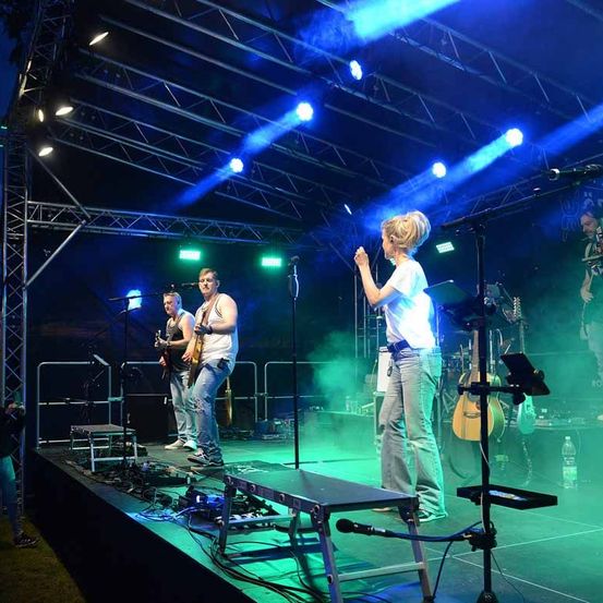 Eine Band tritt auf der Bühne mit blauen und grünen Lichtern auf. Eine Frau singt, während zwei Männer Gitarre spielen. Musikinstrumente und Mikrofone sind auf der Bühne aufgebaut.