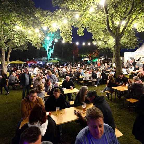 Eine große Menschenmenge versammelt sich in einem Freiluftbereich, möglicherweise bei einem Festival. Sie sitzen an Tischen unter einem Vordach mit Lichterketten. Einige von ihnen halten Gläser und unterhalten sich. Der Bereich ist von Bäumen umgeben. In der Ferne befinden sich Zelte und ein Baum mit grünen Lichtern.