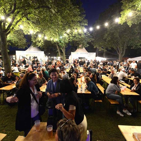 Ein nächtliches Festival unter Bäumen mit Lichterketten, mit zahlreichen Menschen an Tischen sitzend, einigen stehend und anderen im Gespräch, mit Getränken und Essen sichtbar.