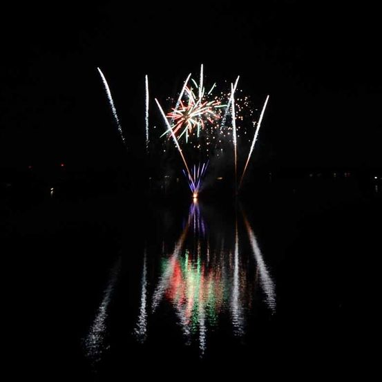 Feuerwerk-Display in der Nacht über einem dunklen Gewässer, das leuchtende Farben reflektiert.