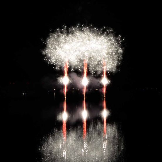 Feuerwerk explodiert in der Nacht über einer dunklen Wasserfläche und spiegelt sich im Wasser darunter.