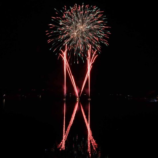 Ein leuchtendes Feuerwerk explodiert in der Nacht, färbt den Himmel mit roten und grünen Schattierungen und spiegelt sich wunderschön auf der ruhigen Wasseroberfläche wider.