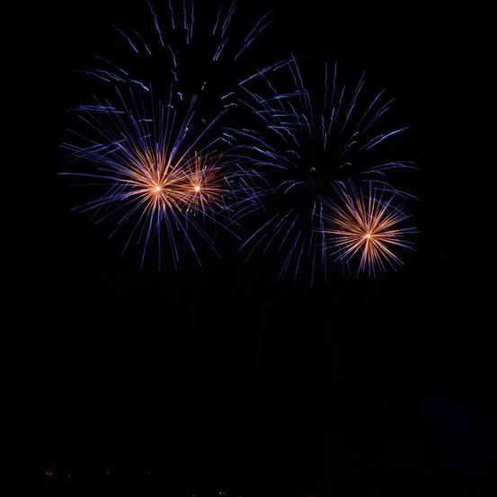 Eine Nachtszene mit Feuerwerk am Himmel. Drei Feuerwerke sind sichtbar, jedes mit blauen und orangefarbenen Farben.