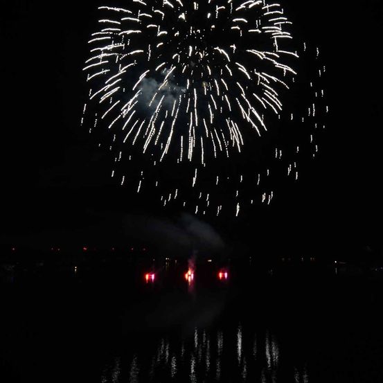 Ein nächtliches Feuerwerk explodiert über einem See, wobei rote Lichter die Wasseroberfläche beleuchten.