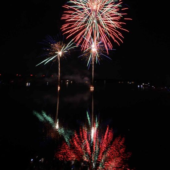 Ein nächtliches Feuerwerksdisplay ist in vollem Gange über einem Gewässer, mit roten, grünen und blauen Farben, die den Himmel erhellen.