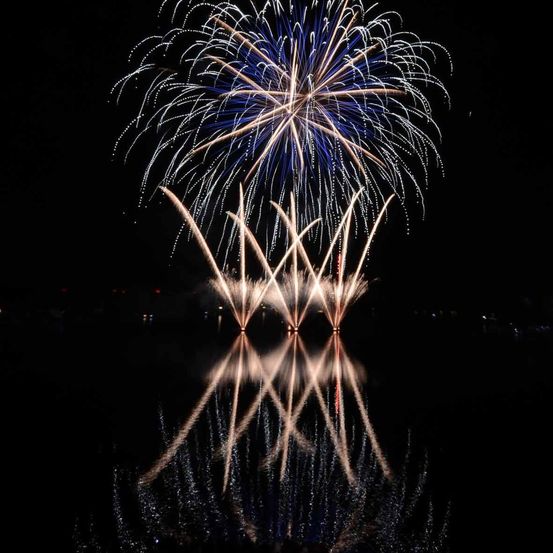 Ein Feuerwerk wird nachts über einer Wasserfläche gezündet, wobei die Reflexion auf der Wasseroberfläche sichtbar ist.
