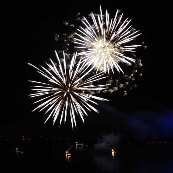 Zwei Feuerwerke explodieren in der Nacht und verbreiten Funken und Licht. Der Hintergrund ist dunkel, und es gibt schwache Lichter von der Stadt und Gebäuden.