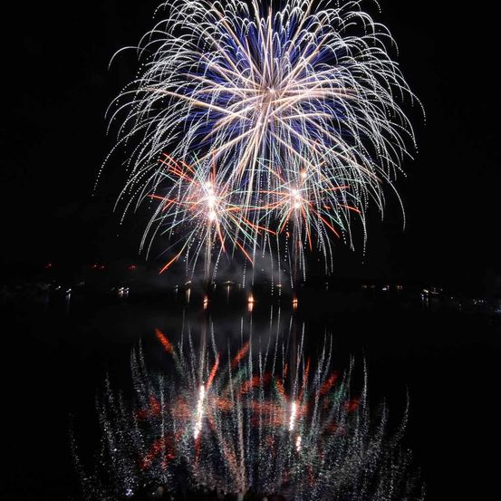 Ein Nachthimmel voller bunter Feuerwerke, die über einem ruhigen See explodieren. Die Reflexion der Feuerwerke ist auf dem Wasser zu sehen.