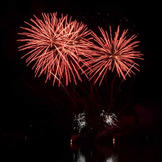 Zwei rote Feuerwerke explodieren in der Nacht über einer ruhigen Wasserfläche und reflektieren Licht.
