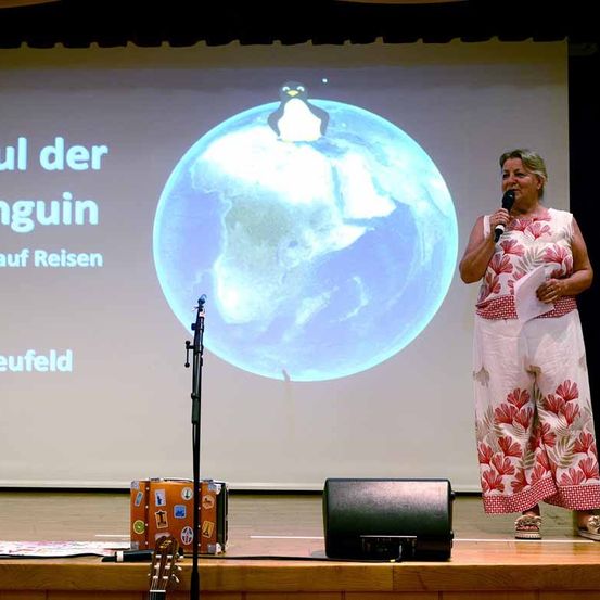Eine Frau auf der Bühne hält ein Mikrofon und spricht. Hinter ihr zeigt eine Projektionsleinwand eine Erde mit einem Pinguin darauf. Der Text lautet 'ul der guin auf Reisen euelfeld'.