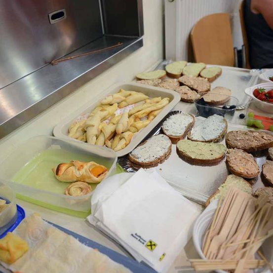 Ein Tisch ist mit verschiedenen Lebensmitteln gefüllt, darunter Sandwiches, Gebäck und Früchte. Es gibt Schalen mit Erdbeeren und Blaubeeren und Utensilien wie Chopsticks. Eine Person ist teilweise im Hintergrund zu sehen.