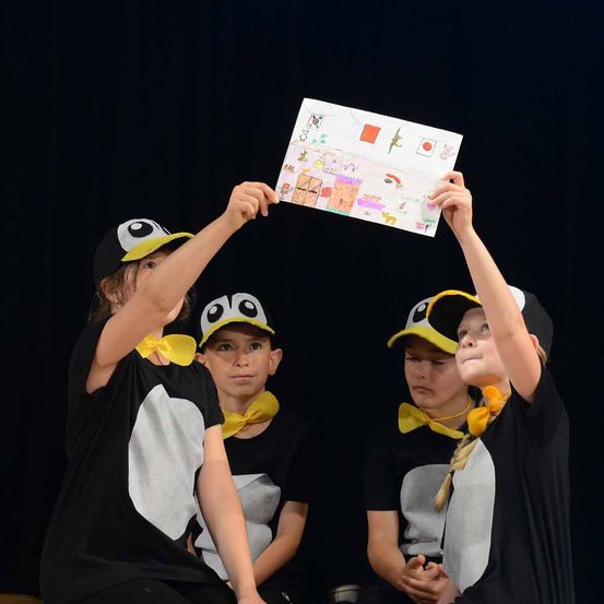 Kinder in Pinguinkostümen halten ein Bild auf der Bühne hoch. Ein Kind hält das Bild mit beiden Händen, während andere zusehen.