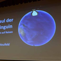 Eine Folie zeigt eine Erde mit einem Pinguin darauf, beschriftet mit 'Paul der Pinguin auf Reisen'. Die Erde ist blau und zeigt die Erde.